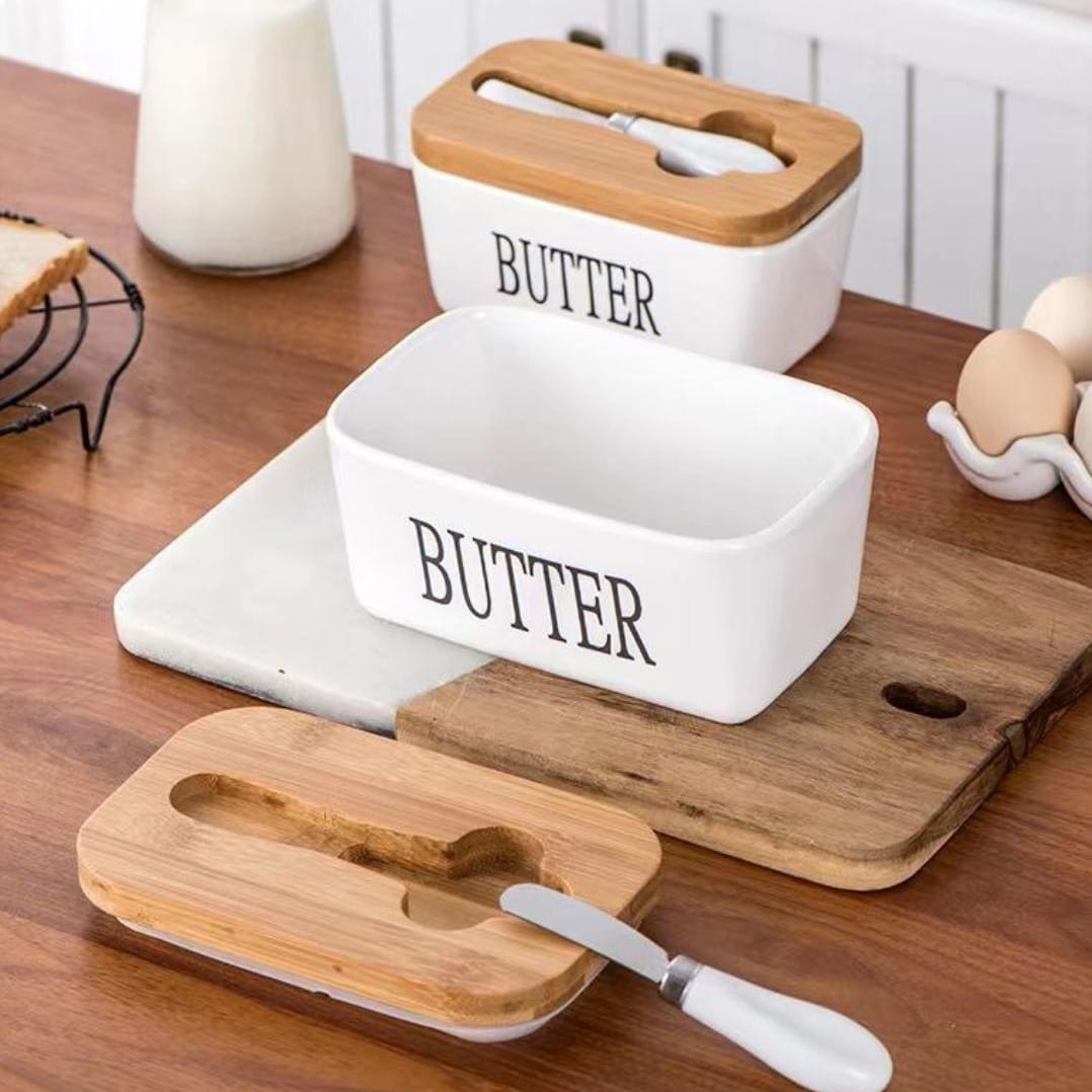 Weisser Butterbehälter mit schwarzer "Butter" Aufschrift neben Holzdeckel, Buttermesser in einem Aufstellbeispiel auf einem Tisch