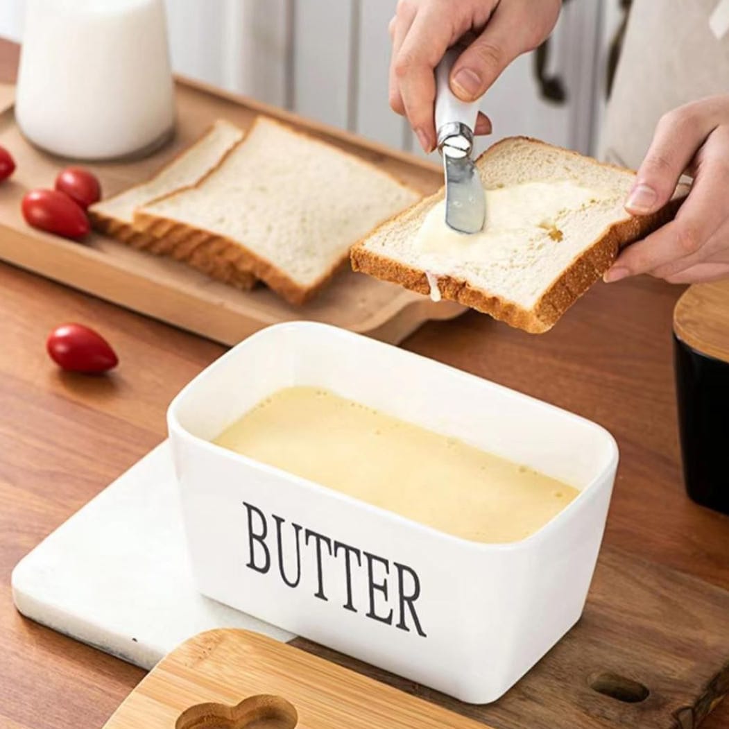 Weisser Butterbehälter mit schwarzer "Butter" Aufschrift neben Holzdeckel, Buttermesser in einem Anwendungsbeispiel