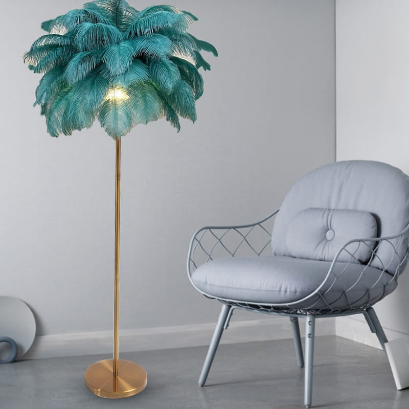 Stehlampe mit blauen Straussenfedern und goldenem Ständer neben grauem Stuhl und grauer Wand