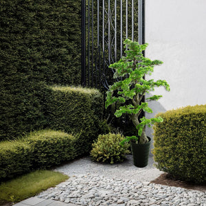 Künstlicher, detaillierter Bonsai mit schwarzem Topf in einem Aufstellbeispiel neben Gebüsch und Steinboden