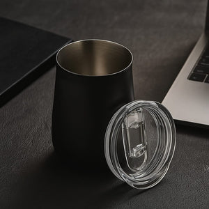 Schwarzer Thermobecher mit transparentem Deckel entfernt auf einem Tisch mit Laptop im Hintergrund