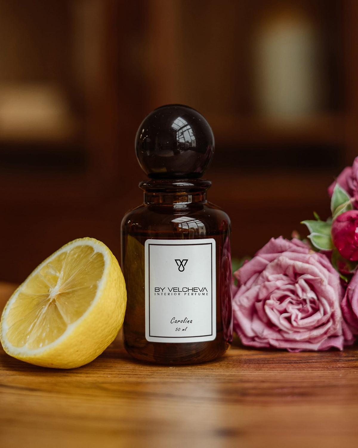 Raumduft Spray in kleiner, braunen Glasflasche mit schwarzem Deckel neben einer halben Zitrone und einer pinken Rose